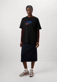 Camiseta de algodón negra con el texto "LIONESSES" en azul y rojo, combinada con una falda larga de mezclilla y zapatillas blancas. Presenta mangas cortas y un corte relajado.