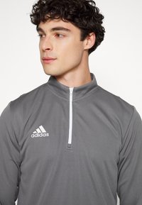 Grijze kwart-zip pullover van lichtgewicht materiaal, met een witte rits en een geborduurd wit Adidas-logo op de linkerkant van de borst.