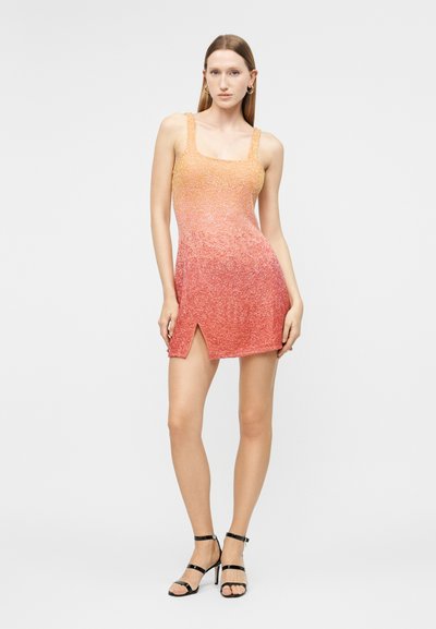 Γυναίκα που φοράει αμάνικο μίνι φόρεμα με διαβάθμιση από πορτοκαλί σε κοραλί, με σκίσιμο μπροστά στον μηρό και μαύρα ψηλοτάκουνα σανδάλια με λεπτά λουριά.