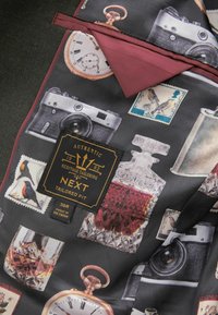 Silk tie featuring a dark background with vintage camera, clock, and bottle illustrations. Burgundy accent at the collar and label visible.