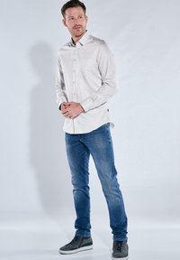 Helles graues Button-Up-Hemd mit langen Ärmeln, kombiniert mit blauen Jeans und grauen Sneakers. Verfügt über eine einzelne Brusttasche.