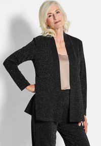 Zwarte sprankelende blazer met een losse pasvorm en een open voorkant; lange mouwen en een zacht beige top eronder. Soepele textuur en elegante drape.