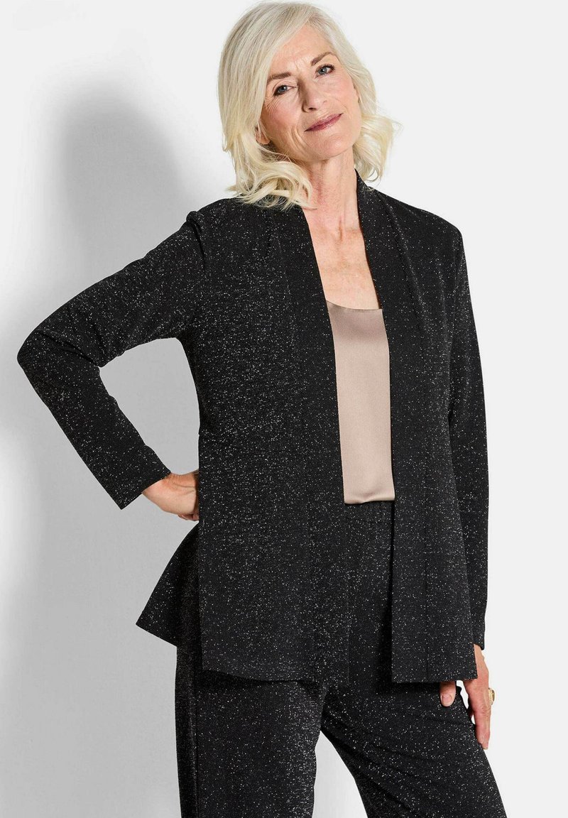Zwarte sprankelende blazer met een losse pasvorm en een open voorkant; lange mouwen en een zacht beige top eronder. Soepele textuur en elegante drape.