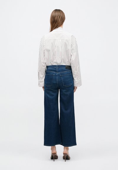 Világos színű csíkos ing, sötétkék, bőszárú farmerrel párosítva. A farmer hátoldalán zseb található, fekete derékpántos címkével.