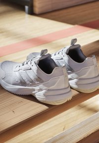 Graue Sportschuhe mit einer strukturierten Oberseite, versehen mit drei weißen Streifen, einer gepolsterten Sohle und einer Schlaufe an der Ferse für einfaches An- und Ausziehen.