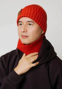 Czerwona dziana czapka o prążkowanej fakturze, zestawiona z pasującym czerwonym kominem. Model nosi czarną bluzę z kapturem. Czapka ozdobiona skórzanym akcentem.