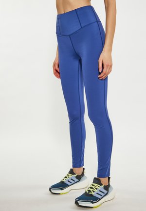 Leggings bleus taille haute avec une texture lisse et extensible, présentant des coutures contrastées. Assortis à des chaussures de sport noires et bleues.
