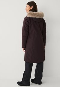 Bordeaux lange jas met furry capuchon, waterafstotende stof, relaxed fit en twee zijzakken, gecombineerd met donkere jeans en zwarte schoenen.