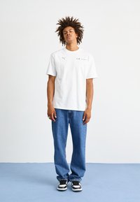 Balta īsām piedurknēm T-kreklu ar Puma logo un Motorsport detaļām, apvienojot ar zilu platā stila džinsu un melniem un baltiem kedām.