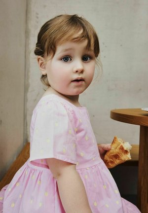 Jong kind met lichtbruin haar in een roze jurk, zittend naast een houten tafel binnenshuis, terwijl het een croissant vasthoudt en eet.