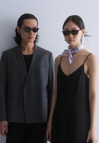 Dos personas están de pie una al lado de la otra. El hombre lleva un blazer gris de doble botonadura, una camisa negra y gafas de sol. La mujer viste un vestido negro de tirantes y una bufanda de un color claro.
