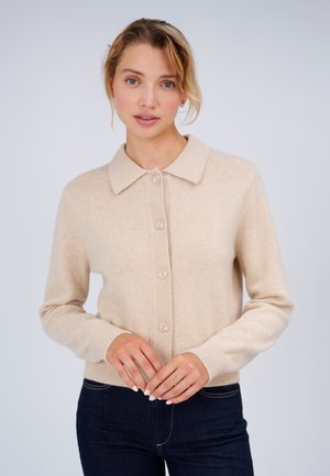 Frau mit hellem Haar, die eine beige, knöpfbare Strickjacke und dunkle Jeans trägt, steht vor einem einfarbigen hellen Hintergrund.
