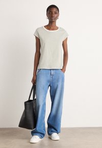 Maglietta a maniche corte a righe in crema e oliva, abbinata a jeans blu larghi. Modella che tiene una borsa a tracolla nera, sneakers bianche. Sfondo neutro.