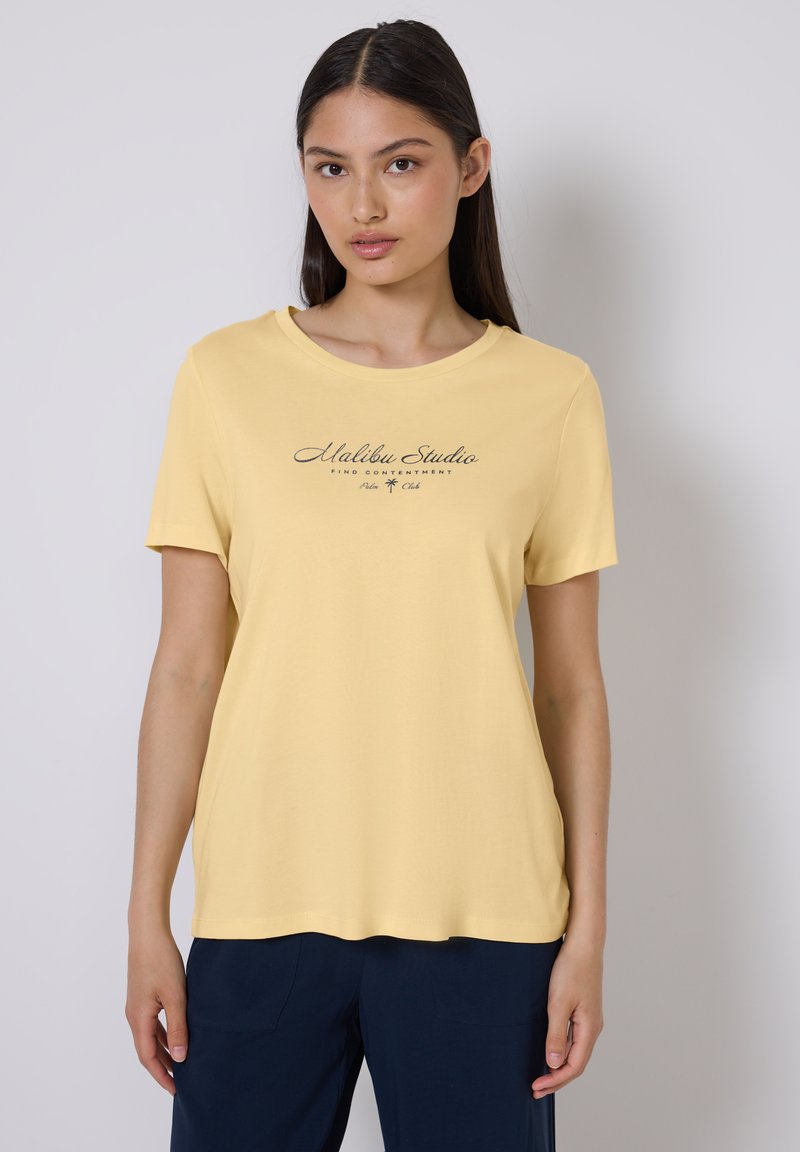 T-shirt en coton jaune à manches courtes, avec un col rond et un graphique texte à l'avant portant la mention "Malibu Studio" en impression foncée.