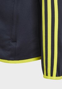 Giacca sportiva blu navy con tre strisce gialle lungo le maniche, tessuto strutturato e una rifinitura gialla a contrasto sull'orlo e sui polsini.