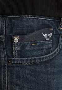 Jean en denim bleu foncé avec une petite poche avant contenant une étiquette noire avec un logo ailé blanc. Couture et rivets visibles.