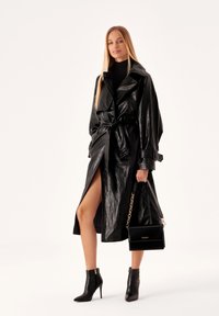 Musta, kiiltävä trench coat, jossa on sidottava vyötärö, sivureikä ja leveä kaulus; yhdistetty korkeakorkoisiin nilkkasaappaisiin ja pieneen mustaan käsilaukkuun.