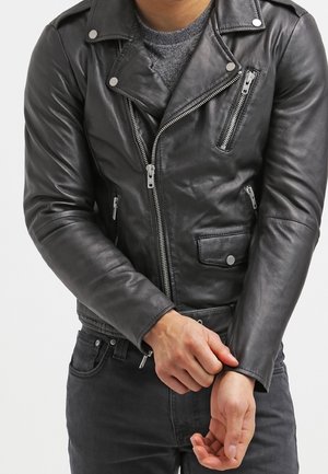 Personne portant une veste de motard en cuir noir avec des fermetures éclair et des boutons argentés, ajustant la manche gauche par-dessus une chemise grise et un pantalon noir.