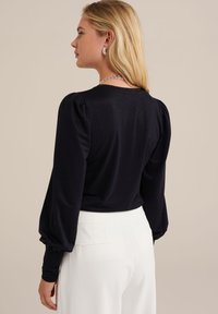 Melnā gar piedurkņu blūze ar uzpūstām plecu daļām, apaļu kakla izgriezumu un gludu tekstūru, apvienota ar baltām piegriezuma biksēm.