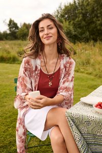 Jeune femme aux longs cheveux assise sur une chaise verte en plein air, tenant une tasse, portant un haut rouge, une jupe blanche, un châle à motifs et des boucles d’oreilles en forme de feuille.