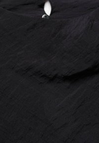 Zwart kledingstuk van een geweven stof met een lichte glans. Ronde kraag met een sleutelgatopening en knoopdetail op de achterkant.