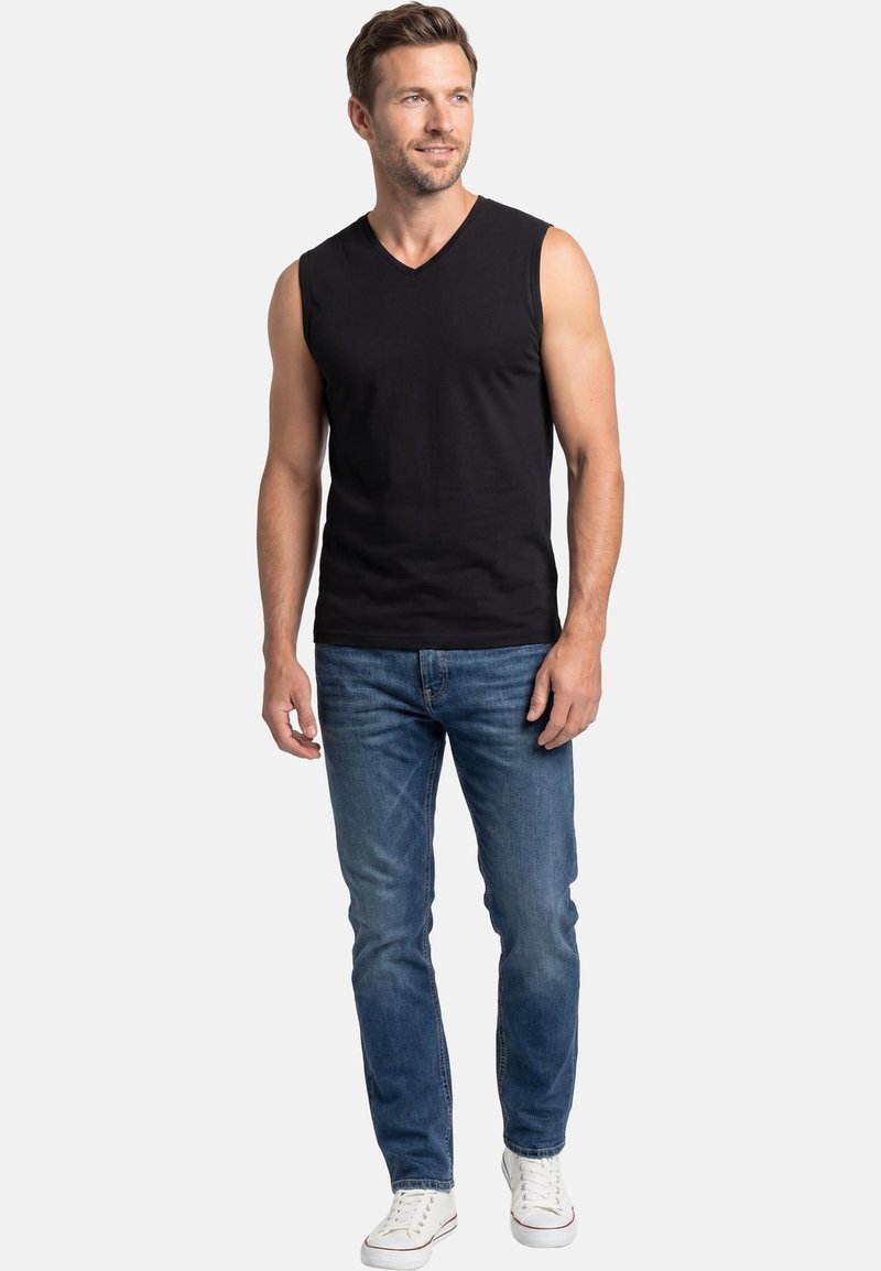 Mężczyzna ubrany w czarną bezrękawnikową koszulkę z dekoltem w serek, niebieskie dżinsy i białe trampki, stojący na tle jasnego, jednolitego tła, patrzący lekko w prawo.