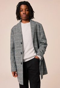 Cappotto grigio a quadri con revers a punta, design monopetto e bottoni grandi, indossato sopra un maglione a costine grigio chiaro e pantaloni neri.