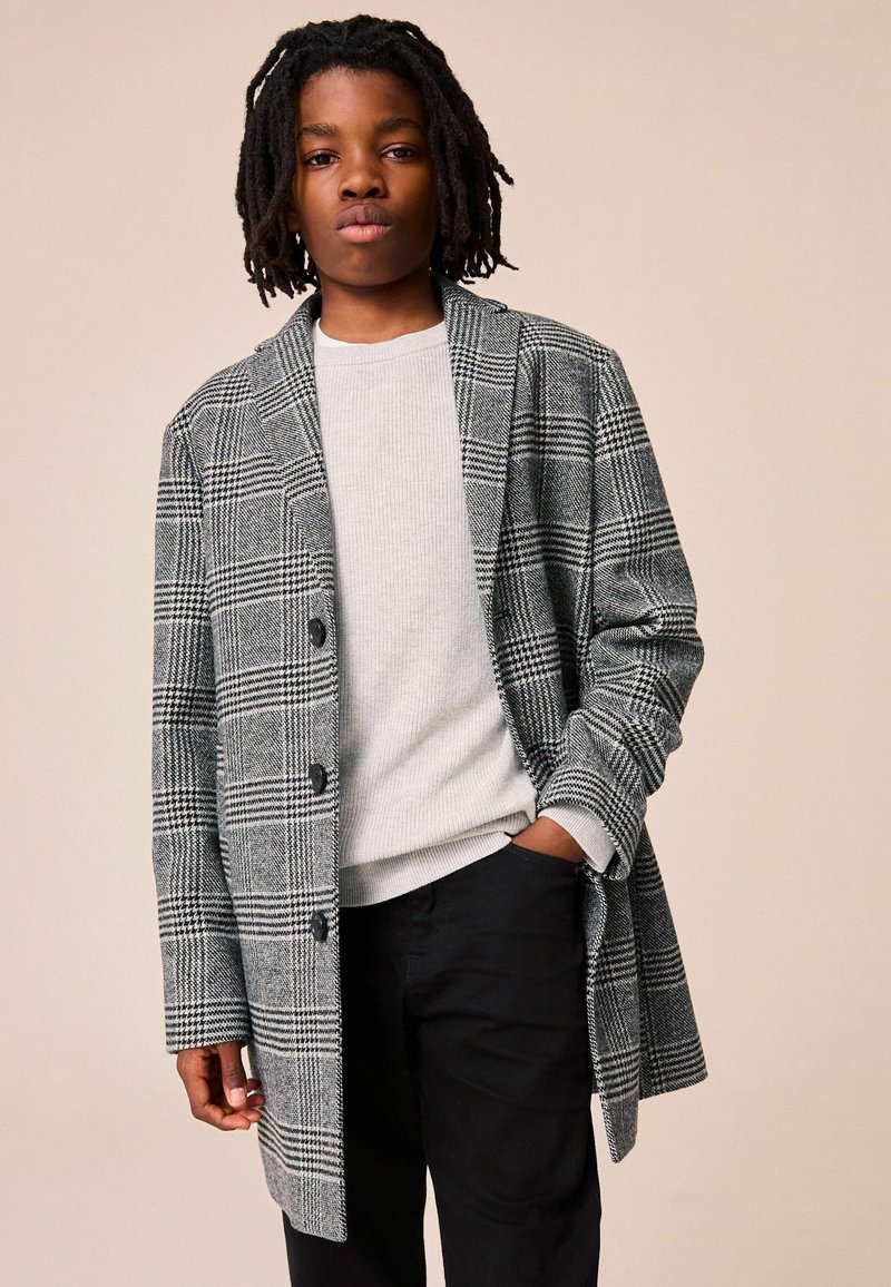 Cappotto grigio a quadri con revers a punta, design monopetto e bottoni grandi, indossato sopra un maglione a costine grigio chiaro e pantaloni neri.