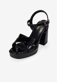 Sandalias de plataforma de charol negro con un tacón grueso, que cuentan con correas entrelazadas y una correa en el tobillo con una hebilla en tono dorado.