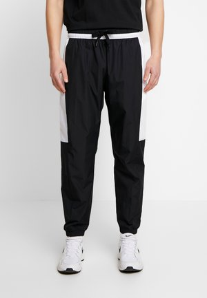 Homme portant un pantalon de survêtement noir et blanc avec des poignets élastiques, une taille à cordon et des baskets blanches avec des détails noirs.