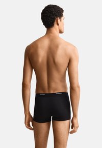 Giovane uomo con capelli corti e ricci che indossa boxer neri GANT, mostrato di spalle su uno sfondo chiaro e uniforme.