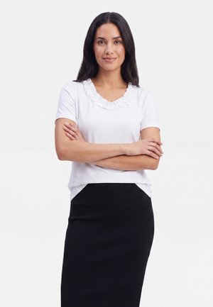 Žena s dlouhými tmavými vlasy v bílém krátkém tričku s nařaseným límečkem a černé sukni stojí se zkříženýma rukama.