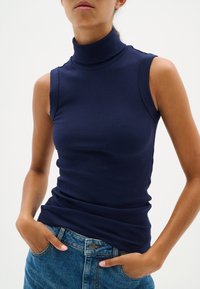 Top senza maniche di colore blu navy con texture a costine. Design aderente con finitura liscia e colletto alto. Abbinato a un paio di jeans in denim.