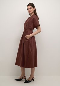Brown, knee-length dress with short sleeves, belted waist, and side pockets. Smooth fabric and a slightly flared silhouette. Black pointed shoes.