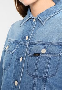 Ljusblå denimjacka med en strukturerad krage, silverfärgade tryckknappar och två bröstfickor med Lee-logotypen på en svart lapp.