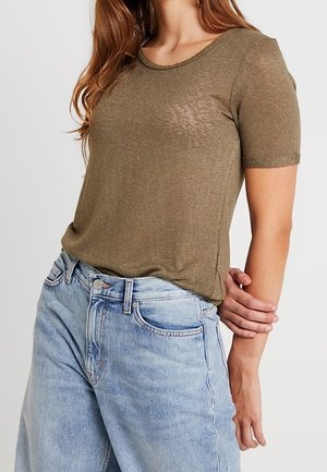 Femme portant un t-shirt court vert olive rentré dans un jean en denim bleu clair taille haute, une main tenant le bras opposé.