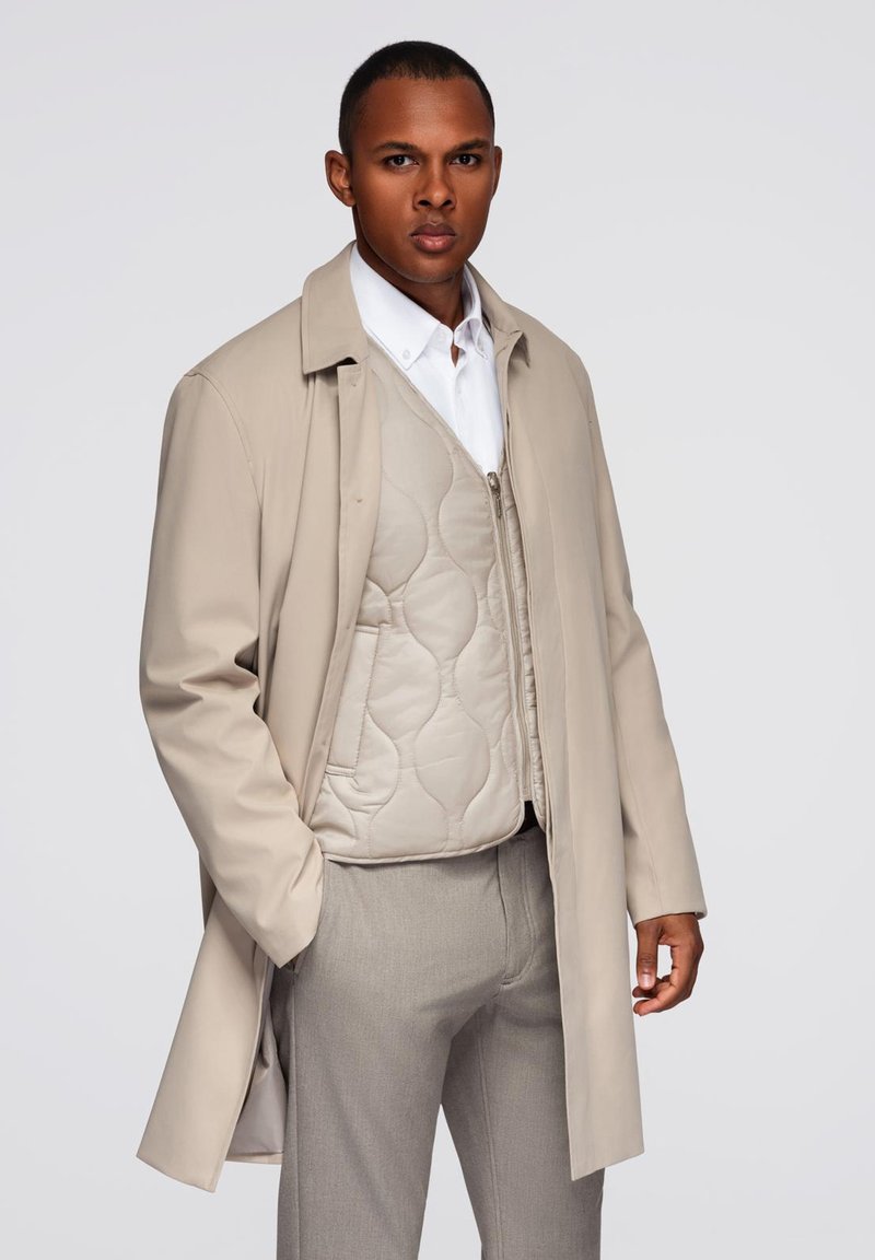 Cappotto beige con maniche lunghe, abbinato a un gilet beige trapuntato con chiusura a zip. I pantaloni grigi completano l'outfit.