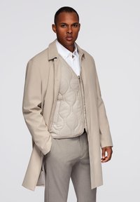 Abrigo beige de manga larga, combinado con un chaleco beige acolchado con cierre de cremallera. Pantalones grises completan el conjunto.