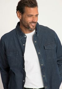 Camicia in denim blu scuro con abbottonatura frontale, due tasche sul petto e colletto. Indossata sopra una maglietta bianca, con una finitura liscia e testurizzata.