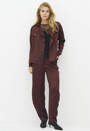 Woman wearing dark burgundy loose jacket and matching pants with a black lace top and black high-heeled shoes standing against a white wall.