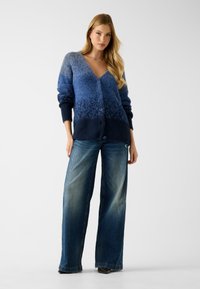 Gebreid blauwe vest met een verloop van licht naar donker, voorzien van een V-hals, knoopsluiting aan de voorkant en geribbelde manchetten, gecombineerd met wijde jeans.