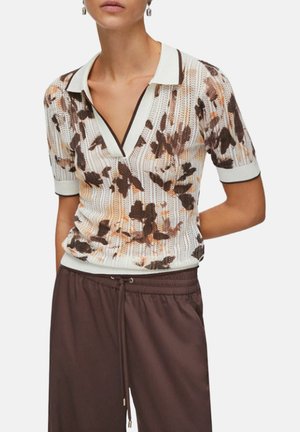 Kvinde iført en hvid strikket top med korte ærmer med brune blomsterprint og brune bukser med snøre, vist fra nakke til hofter.