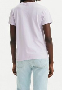 Persona con piel oscura que lleva una camisa de manga corta de color púrpura claro y jeans de color azul claro, vista desde la parte de atrás contra un fondo blanco.