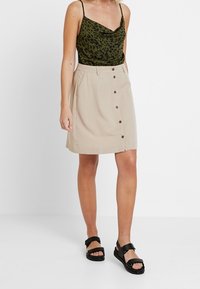 Grünes Oberteil mit Camouflage-Muster und dünnen Trägern, kombiniert mit einem beigen Rock mit Frontknöpfen. Schwarze Sandalen mit breiten Riemen.