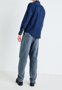 Camisa de mezclilla azul oscuro con cuello abotonado, mangas largas y dobladillo curvado, combinada con pantalones grises claros de corte holgado y zapatos negros.