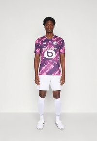 Camisola de futebol com padrão roxo e rosa, gola redonda, calções brancos e meias brancas. Apresenta um logótipo e texto de patrocinador na parte da frente.