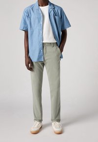 Kortsleeve blå chambrayskjorta över en vit t-shirt, parat med ljusgröna byxor och vita sneakers med bruna sula.