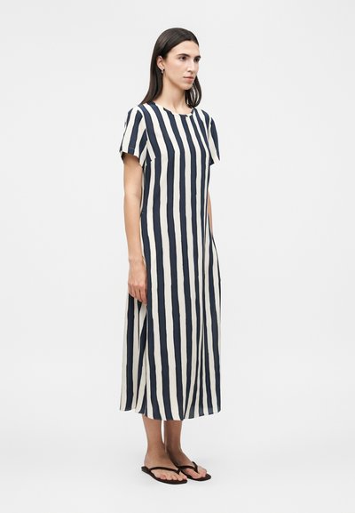 Γυναίκα που στέκεται φορώντας μαύρο και λευκό midi φόρεμα με κάθετες ρίγες και κοντά μανίκια, και μαύρα σανδάλια τύπου φλιπ-φλοπ, στραμμένη ελαφρώς προς τα δεξιά.