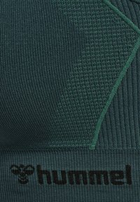 Dunkelgrünes Sportoberteil aus strukturiertem Rippgewebe. Es hat ein geometrisches Design und das schwarze 'hummel'-Logo am unteren Ende.