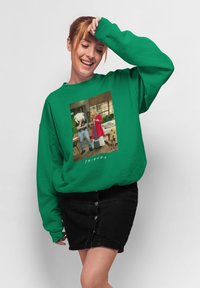 Maglione di felpa verde oversize con una scena stampata di un programma televisivo, con scollo rotondo e vestibilità morbida. Abbinato a una gonna nera con bottoni anteriori.